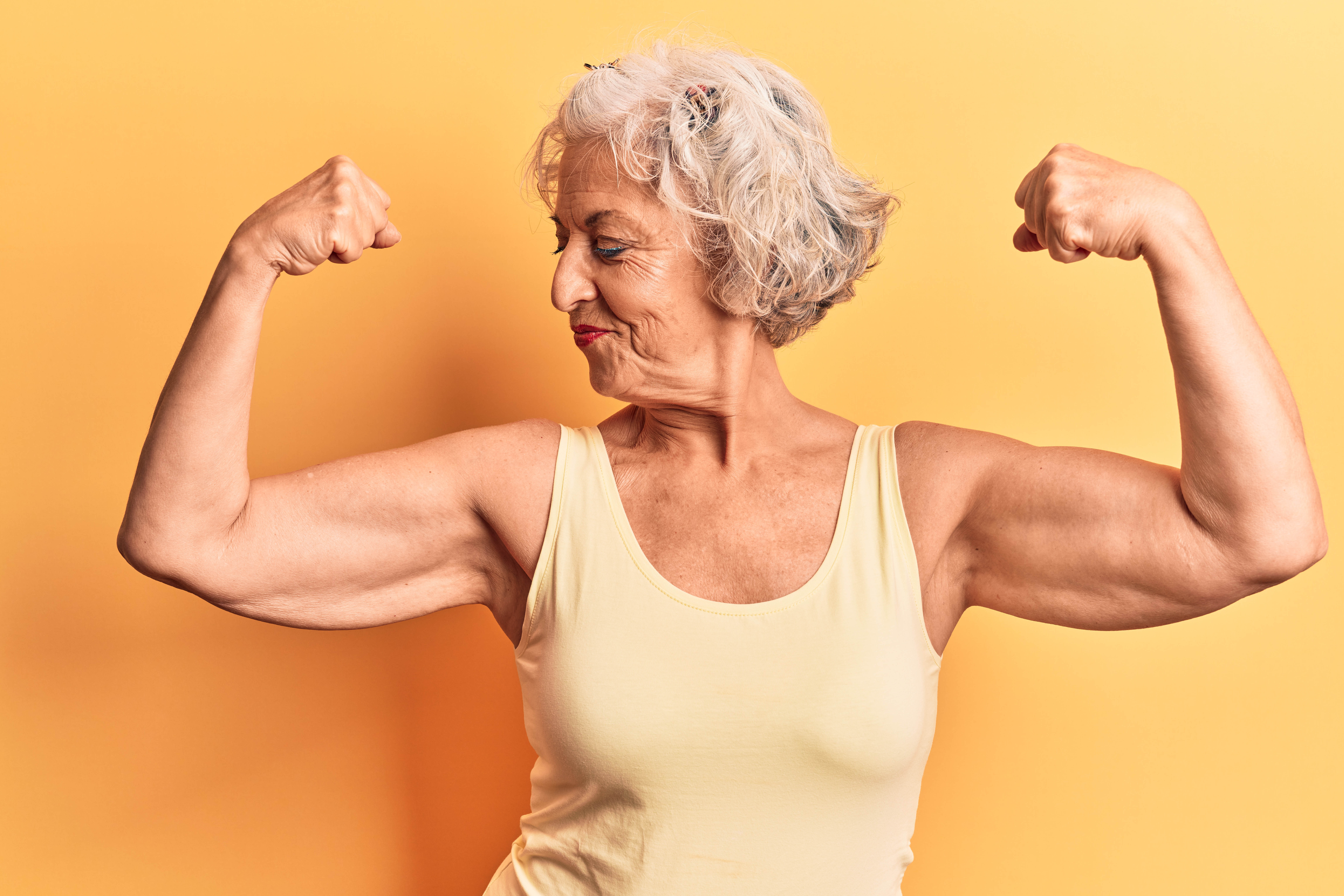 Older woman flexing and looking sideways at her bicep on a yellow background